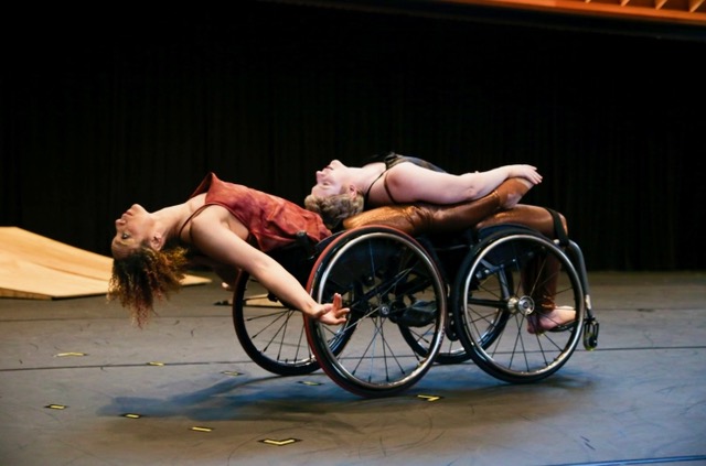 Alice Sheppard and Laurel Lawson lay backwards in their wheelchairs, torsos and throats open. Alice is a multiracial Black woman with coffee-colored skin and short curly hair, and Laurel is a white woman with cropped blonde hair. They wear shimmery costumes in autumnal tones. A small set of wood ramps appear in the background. Photo by Noor Eemaan Jaffery/ Jacob's Pillow.